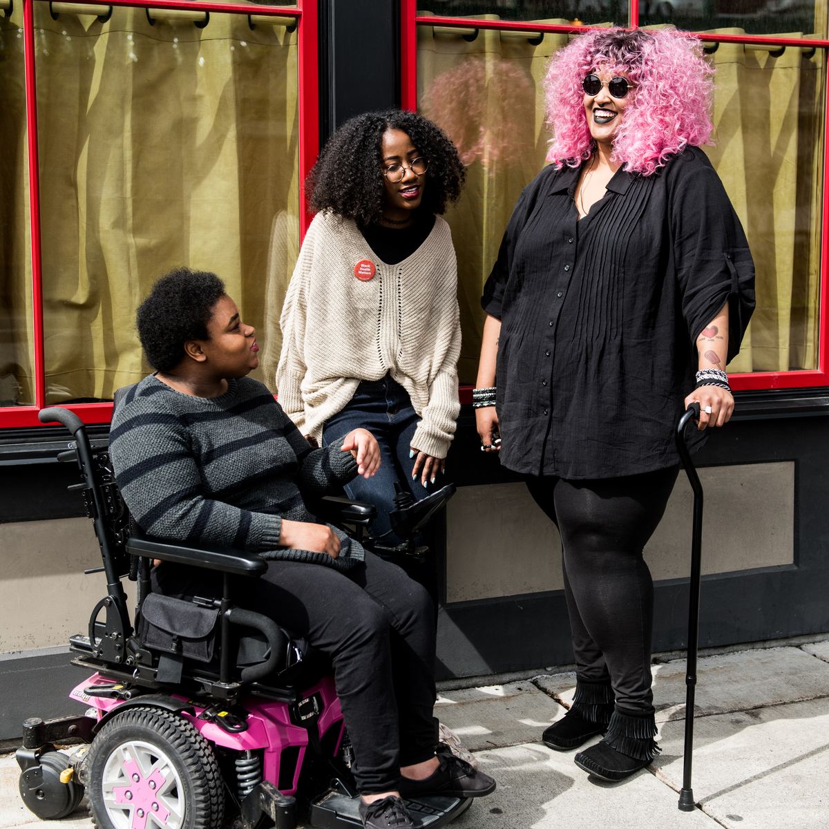 Three friends talking outside, one using a power wheelchair and one a cane